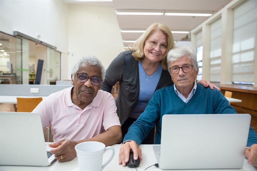 Three older adults smiling around laptops in a bright study room